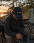 Person sitting by a campfire with a Scclina cap, camping setup, and sunset over water.