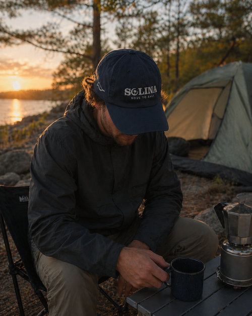 Person sitting by a campfire with a Scclina cap, camping setup, and sunset over water.
